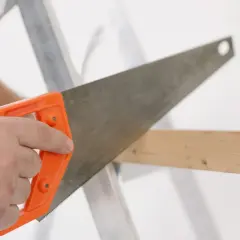 Main tenant une scie à bois à manche orange, coupant une planche. La différence scie à bois scie à métaux est dans la denture.