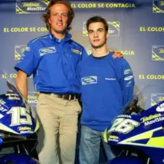 Sete Gibernau, con su edad, posa junto a un joven piloto, ambos con ropa de equipo azul, frente a motocicletas de competición.