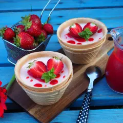 Panna cotta w wafelkach z truskawkami i miętą, obok dzbanuszek z musem truskawkowym.
