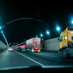 W tunelu panuje noc, światła drogowe oświetlają jezdnię. Samochody i ciężarówki poruszają się po pasach. Widać znak zakazu wyprzedzania dla ciężarówek.
