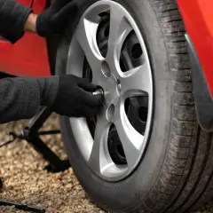 Ręce w rękawiczkach dokręcają śrubę koła czerwonego samochodu. W tle podnośnik i klucz, pomocne przy zmianie opon. To część przygotowań do sezonu, ważna dla rankingu opon letnich.