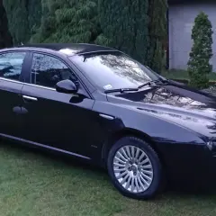 Elegancka, czarna Alfa Romeo 159 stoi na trawniku, otoczona zielenią.