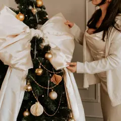 Elegancka stylizacja na święta bożego narodzenia: kobieta w satynowej sukience i swetrze poprawia dużą, kremową kokardę na choince ozdobionej złotymi bombkami.