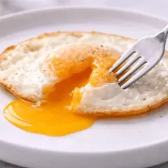 Un œuf miroir, le jaune coulant comme un soleil liquide, est percé par une fourchette sur une assiette blanche.