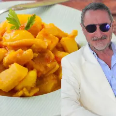 Un hombre con gafas de sol y traje blanco sonríe junto a un plato de papas con choco de la abuela, un guiso casero.