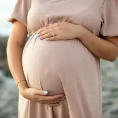 Brzuch kobiety w ciąży, delikatnie otulony beżową sukienką. Dłonie z jasnoniebieskim lakierem na paznokciach spoczywają na brzuchu. Warto pamiętać o bezpieczeństwie, np. unikając retinolu w ciąży.