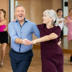 Uśmiechnięta para seniorów w rytmie tańca na 3. W tle inne pary ćwiczą kroki.