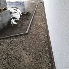 Ułożono kruszywo kamienne tłuczeń wzdłuż białego muru, obok palet z materiałami budowlanymi.