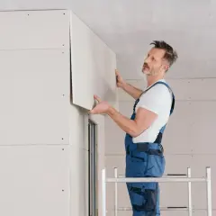 Operaio in tuta blu posiziona un pannello di cartongesso sul soffitto, completando il lavoro di fissare cartongesso direttamente su parete.