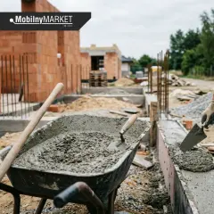Budowa fundamentów: w taczce świeży beton, a pracownik ręką z kielnią układa go w szalunku. Jaki cement na fundament? Tu widać pracę z betonem.