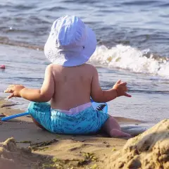 Niemowlę w kapeluszu i kąpielówkach z rekinami siedzi na plaży, ciesząc się wakacjami z niemowlakiem nad morzem.