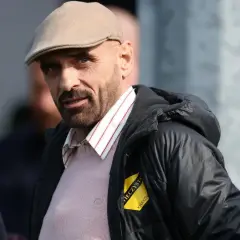 Mężczyzna w bejsbolówce i kamizelce z naszywką "Wieczysta Kraków właściciel" spogląda w obiektyw.