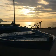 Un'imbarcazione blu riposa sulla riva al tramonto, con il sole che filtra tra le nuvole e illumina il mare calmo.