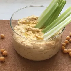 Domowy hummus przepis: kremowa pasta z ciecierzycy w szklanej misce, podana z łodygami selera i ziarnami ciecierzycy.