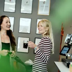 Två kvinnor, en grafisk formgivare, pratar vid en kaffemaskin. Väggen bakom dem är prydd med diplom.