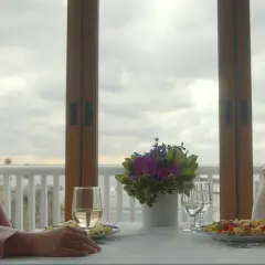 Dwie kobiety siedzą przy stole, patrząc na siebie. Jedna w różowej marynarce, druga w białym golfie. W tle okno z widokiem na pochmurne niebo. Niech spoczywaj w pokoju.