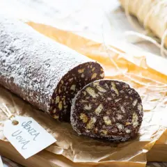 Salame al cioccolato senza uova, cosparso di zucchero a velo, con una fetta tagliata che mostra i biscotti sbriciolati.