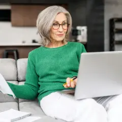 Femme âgée souriante, assise sur un canapé, travaillant sur son ordinateur portable et consultant des documents. Elle semble gérer son **apa à domicile montant**.