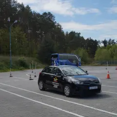 Na placu manewrowym, gdzie obowiązuje ruch lewostronny, czarny samochód szkoleniowy i czerwony pojazd czekają na kursantów. W tle niebieski autokar.
