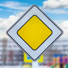 Znak droga z pierwszeństwem, biały romb z żółtym środkiem, na tle błękitnego nieba z chmurami.