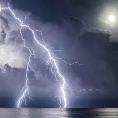 Dramatic lightning photography captures multiple bolts striking the ocean under a stormy sky with a bright moon.