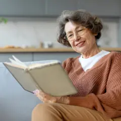 Uśmiechnięta starsza pani czyta książkę. W tle kuchnia. Może to być historia o tym, kto jest właścicielem mieszkania po podpisaniu umowy dożywocia.