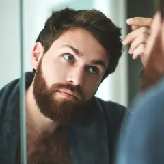 Hombre con barba revisando su pelo graso frente al espejo, con una bata puesta.