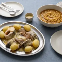 Bullit de peix servido en una bandeja de plata con patatas y una salsa cremosa, acompañado de un cuenco de arroz y un plato vacío.