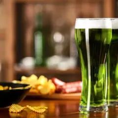 Trzy szklanki zielonego piwa z pianką, obok miska chipsów i kiełbasa. Idealne na świętowanie!