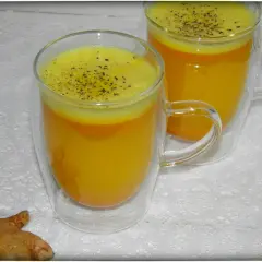 Zwei Gläser mit leuchtend gelbem Kurkuma-Wasser, bestreut mit schwarzem Pfeffer. Daneben liegen frische Kurkumawurzeln. Ein einfaches Rezept für Wohlbefinden.