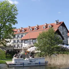 Hotel Anek Mrągowo nad jeziorem, z łodzią przy pomoście i zielenią.