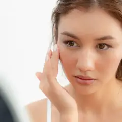 Une jeune femme examine son visage, préoccupée par la sécheresse cutanée. Sa peau semble tendue et manque d'hydratation.
