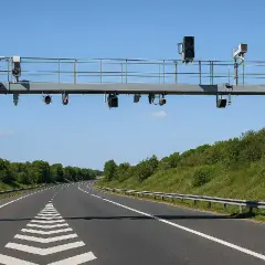 Portique de péage sans barrière sur une autoroute bordée d'arbres. Des caméras et des feux sont installés sur la structure métallique.