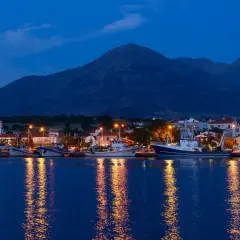 Port z samotraki nocą. Łodzie cumują przy nabrzeżu, a światła odbijają się w spokojnej wodzie. W tle widać góry.