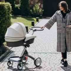 Kobieta w płaszczu prowadzi spacerówkę kubelkową po brukowanej ścieżce, wśród zieleni.