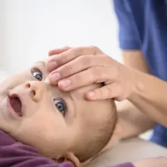 Ein Baby nach Osteopathie, wie ausgewechselt, mit weit geöffnetem Mund und großen Augen, während eine Hand sanft seine Stirn berührt.