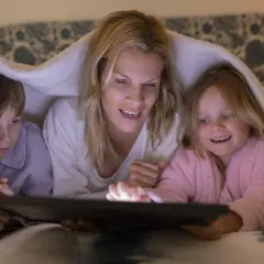 Mama z dziećmi pod kocem ogląda bajkę na tablecie. To piękny przykład, jak wychować dziecko w bliskości i wspólnocie.
