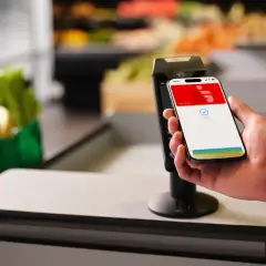 Einkaufen leicht gemacht: So funktioniert Apple Pay. Ein iPhone wird zum Bezahlen an ein Terminal gehalten, im Hintergrund sind Lebensmittel zu sehen.
