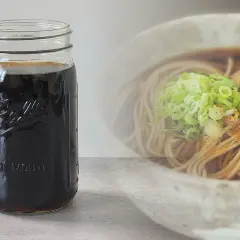 A jar of dark soba dipping sauce sits next to a bowl of soba noodles with green onions. Perfect for your soba dipping sauce recipe.