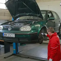 Mechanik w czerwonej kurtce sprawdza podniesiony na podnośniku zielony samochód. To kluczowy moment, by dowiedzieć się, jak dbać o samochód.