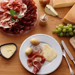 Une table dressée pour une abondance raclette : charcuteries, fromages, pommes de terre, raisins et poêlon de fromage fondu.