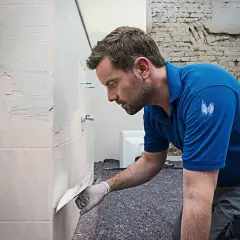 Mann spachtelt Fliesen mit einer Kelle. Er trägt Handschuhe und ein blaues Polohemd.