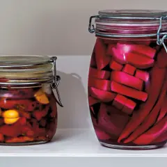 Två glasburkar med inlagd paprika och andra grönsaker. Den större burken innehåller röda, avlånga bitar, troligen inlagd paprika.