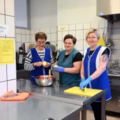 Trzy kobiety w fartuchach obierają jabłka w szkolnej kuchni.