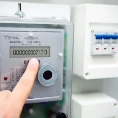 Dito che controlla il contatore luce digitale, mostrando il consumo attuale. Accanto, un quadro elettrico con interruttori.