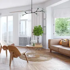 Jasny salon z drewnianą podłogą, kanapą, dwoma fotelami i stolikiem. Przez okna widać zieleń, a na nich zwiewny woal.
