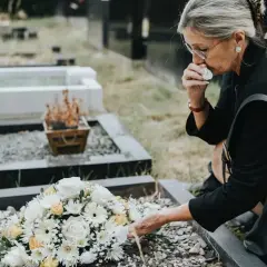 Kobieta w żałobie klęczy przy grobie, ocierając łzy. Zastanawia się, ile wynosi zasiłek pogrzebowy, by móc godnie pożegnać bliską osobę.