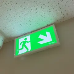 Zielone oświetlenie awaryjne z ikoną biegnącej postaci i strzałką wskazuje drogę ewakuacji. Obok widoczna kamera monitoringu.