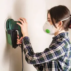 Mujer con mascarilla lijando paredes con una lijadora eléctrica.