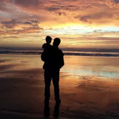 Silhouette di un padre che porta in braccio il figlio sulla spiaggia al tramonto. Un momento di gioia che celebra la festa del papà nel mondo.